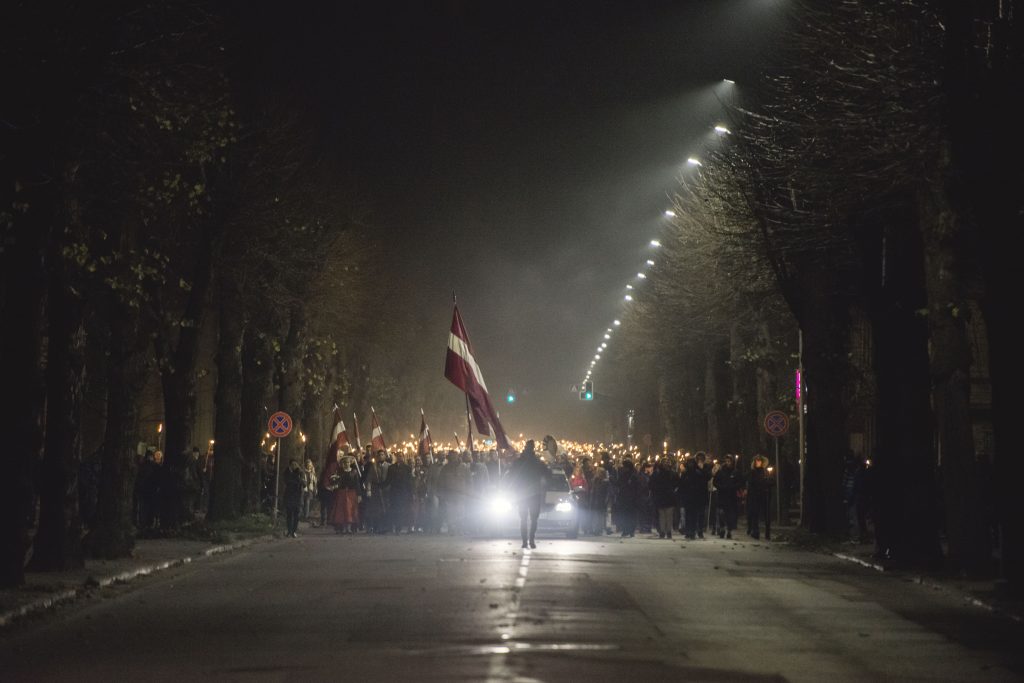 cilvēki ar gaismas lāpām un lielu, plīvojošu Latvijas karogu