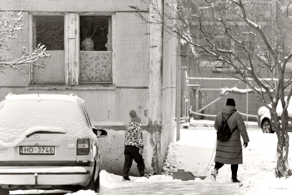 Melnbalta fotogrāfija. Logā aiz aizskariem kungs un uz ielas staigā cilvēki