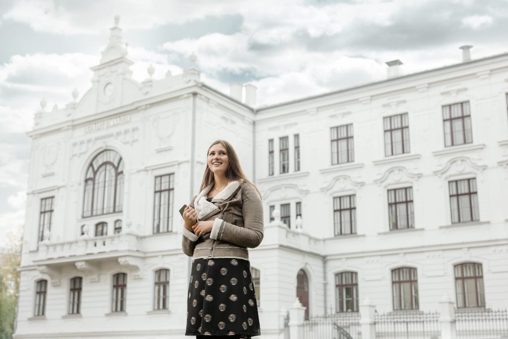 Meitene ar grāmatām rokās, fonā redzama liela, balta ēka