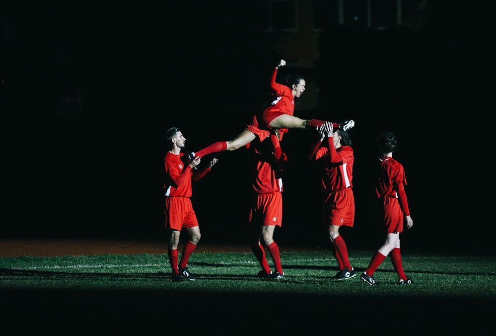 Uz tumša fona pieci futbolisti tērpušies sarkanās formās