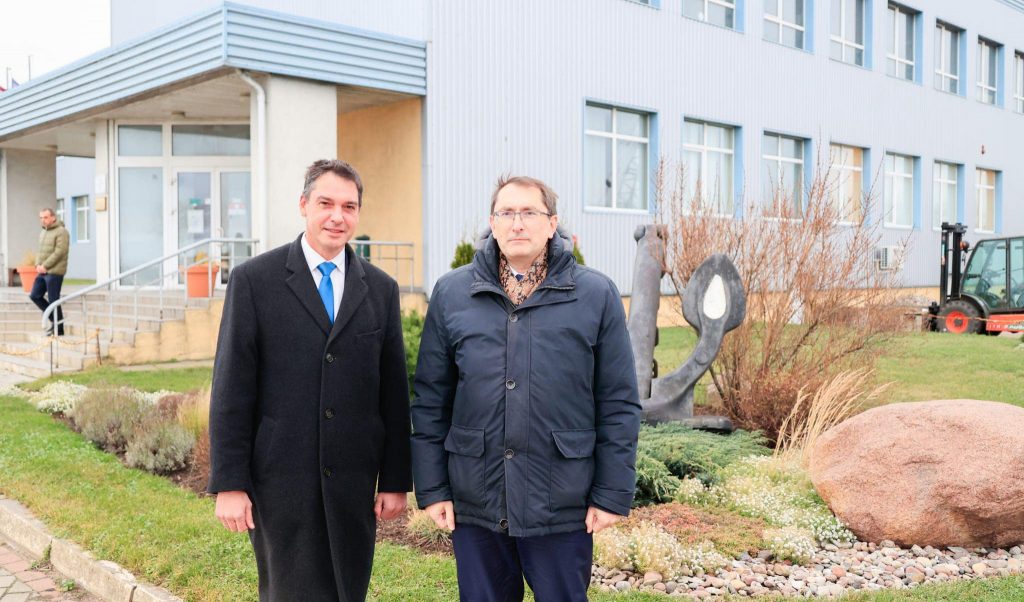 Divi kungi stāv pie baltas ēkas un aiz viņiem novietots pelēks enkurs
