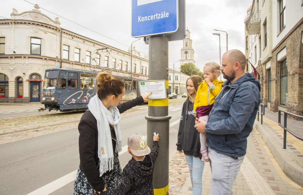 Sabiedriskā transporta pasažieri piestājuši pie pieturas un skata transporta kustības sarakstu