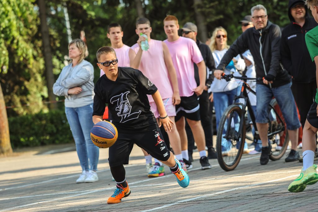 Ielas basketbola sacensības, priekšplānā puisis driblē bumbu, aizmugurē vairāki cilvēki vēro